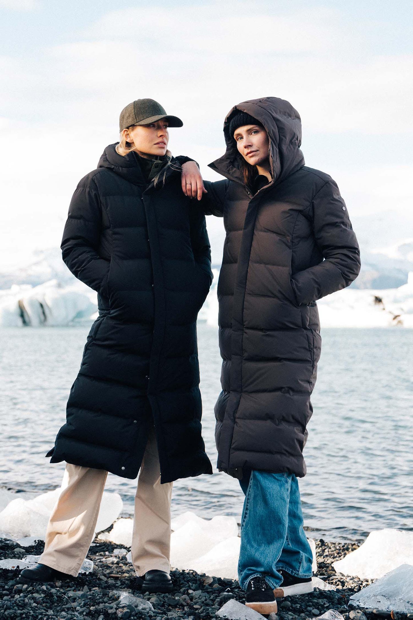 Two people wearing long black puffer coats standing on a rocky shore with water and icebergs in the background.