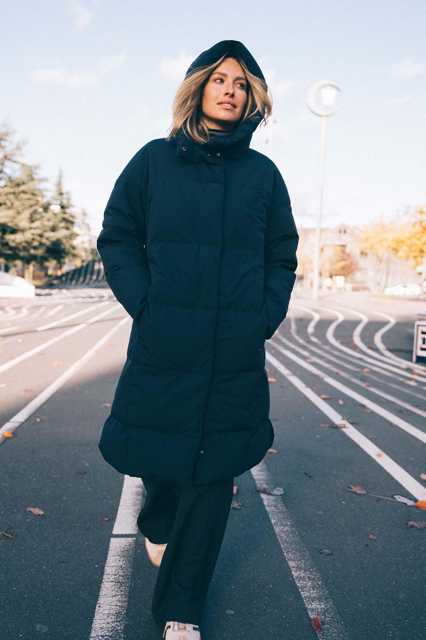 Person wearing a long dark coat standing on a road with trees and buildings in the background